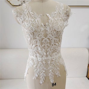 May include: White beaded lace applique with a floral design. The lace is delicate and intricate, with small beads and sequins throughout. The applique is perfect for adding a touch of elegance to any garment.