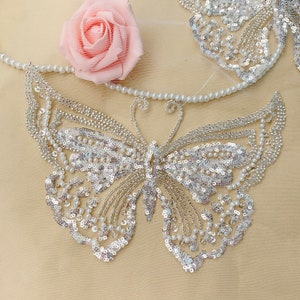 May include: A silver sequined butterfly applique with pearl accents. The butterfly is on a beige fabric background.