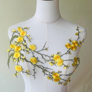 Könnte beinhalten: Gestickte Blumenapplikation mit gelben Blüten und grünen Blättern auf braunen Zweigen. Das Design wird auf einer weißen Schaufensterpuppe präsentiert, die die feinen Details des Stoffes und der Stickerei hervorhebt. Ideal zum Verzieren von Kleidungsstücken.