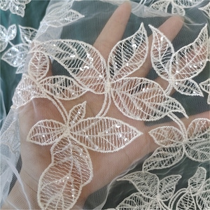 May include: White embroidered lace fabric with sequins. The fabric has a floral design with leaves and a mesh background.