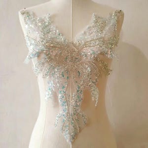 May include: A white lace applique featuring intricate embroidery, silver metallic thread, and iridescent sequins. The design is symmetrical with a V-shaped neckline, showcasing swirling patterns and floral-inspired motifs across the entire surface.