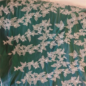 May include: White floral lace fabric on a green background. The lace is made of delicate white leaves and vines. The fabric is sheer and has a soft, flowing texture.