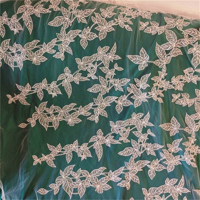 May include: White floral lace fabric on a green background. The lace is made of delicate white leaves and vines. The fabric is sheer and has a soft, flowing texture.