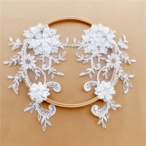 May include: Two white lace appliques with floral designs and 3D flowers. The appliques are displayed on a wooden embroidery hoop.