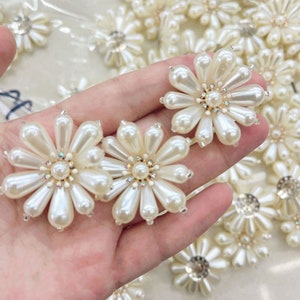 May include: A close-up of a hand holding several white pearl flower embellishments. The flowers have a central rhinestone and are perfect for adding a touch of elegance to clothing or crafts.