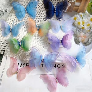Peut inclure: Plusieurs décorations de papillons translucides aux couleurs pastel : bleu, rose, violet, vert et orange. Chaque papillon est orné d'une perle en son centre. Les mots "IMP", "T" et "NGS" sont visibles.