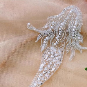 Könnte beinhalten: Eine weiße Perlen-Meerjungfrauen-Applikation mit silbernem Fadenrand. Der Meerjungfrauen-Schwanz ist aus Pailletten und die Haare aus Perlen.