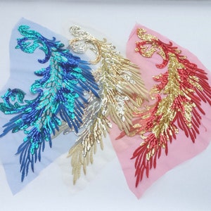 Op de afbeelding: Drie paillettenstoffapplicaties in de vorm van een gestileerde vogel met gouden, rode en blauwe pailletten. De applicaties bevinden zich op een transparante stoffen achtergrond.