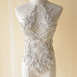 Peut inclure: Une applique blanche en perles et strass avec un motif floral. L'applique est sur un torse de mannequin blanc.