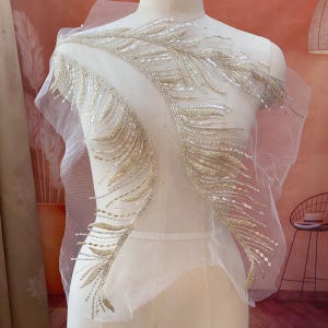 Może przedstawiać: Biała, przezroczysta tkanina z ozdobami z złotych koralików w kształcie piór. Tkanina jest udrapowana na manekinie.