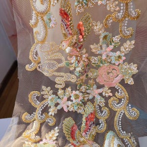 May include: Gold beaded embroidery trim featuring a floral and bird design. The trim incorporates sequins, beads, and pearls in shades of pink, green, and gold.