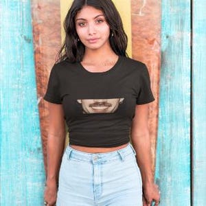 Pu&ograve; includere: Una donna indossa un top corto nero con un'immagine stampata di una bocca e pantaloncini di jeans blu. &Egrave; in piedi davanti a uno sfondo a strisce blu e giallo.