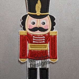 May include: A red and gold glitter embroidered nutcracker with a black hat and a gold trim. The nutcracker has a white face with black eyes, a black mustache, and pink cheeks. The text "Nettielyn Designs" is embroidered on the bottom of the nutcracker.