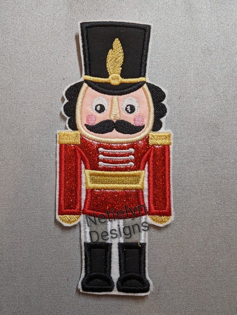 May include: A red and gold glitter embroidered nutcracker with a black hat and a gold trim. The nutcracker has a white face with black eyes, a black mustache, and pink cheeks. The text "Nettielyn Designs" is embroidered on the bottom of the nutcracker.