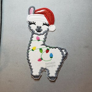 May include: White llama wearing a red Santa hat with white trim and a string of colorful lights around its neck. The llama is standing on a gray surface. The text "Nettielyn Designs" is printed on the llama's back.