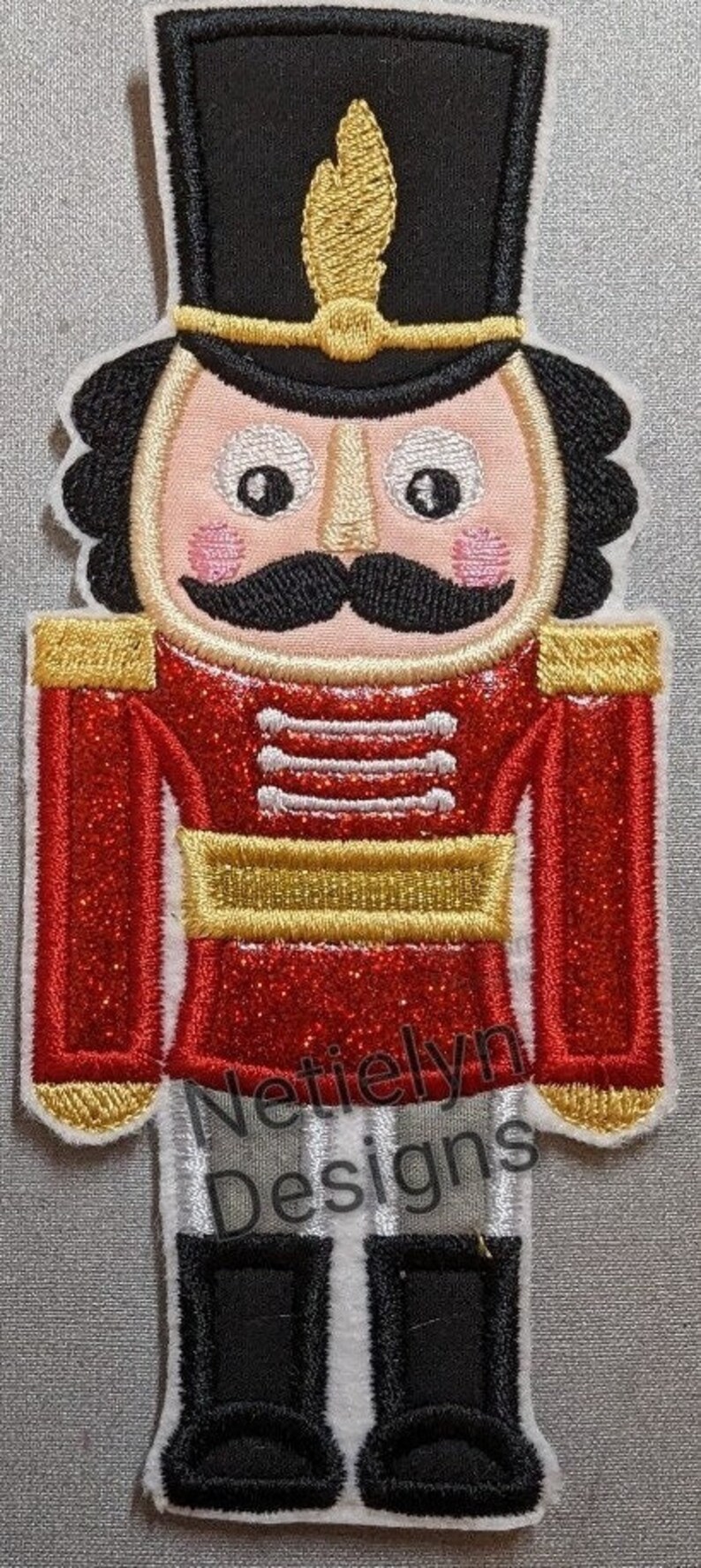 May include: A red and gold glitter nutcracker with a black hat and a white face. The nutcracker has a black mustache and a white beard. The nutcracker is standing on a white background. "Nettielyn Designs" is written in white on the bottom of the nutcracker.