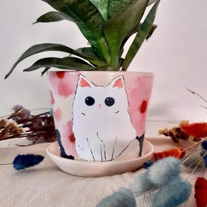 Op de afbeelding: Een witte keramische plantenpot met een roze en rood bloemenontwerp en een witte katillustratie. De pot heeft een bijpassende schotel en is gevuld met een groene slangplant.