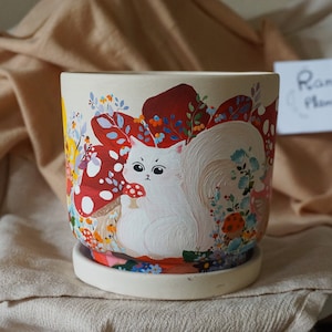 May include: White ceramic planter with a hand-painted design of a white cat sitting in a field of red and white mushrooms and flowers. The planter has a matching saucer.