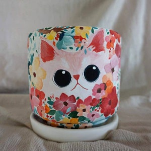 May include: A white ceramic planter with a pink cat face surrounded by colorful flowers. The planter has a white saucer.