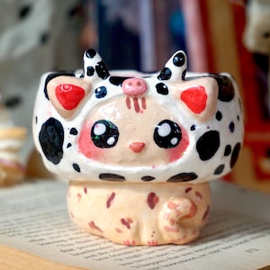 May include: A ceramic planter shaped like a cat wearing a black and white cow costume. The planter has large, black eyes and a pink nose. The cat is sitting on a book.