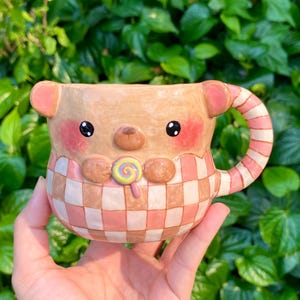 May include: A handmade ceramic mug shaped like a bear, featuring a pink and white checkered pattern. The mug has a bear face with black eyes, pink cheeks, and a lollipop. The handle is striped pink and white.