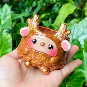 May include: A small, brown ceramic planter shaped like a deer with gold accents and pink ears. The planter has a small opening at the top for planting.