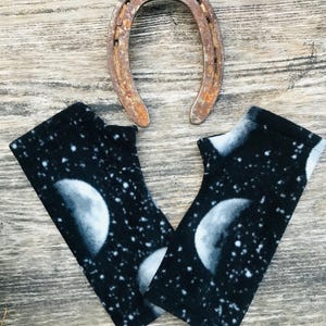 May include: Black fingerless gloves with a celestial moon and star pattern. A rusty horseshoe is positioned above the gloves. The gloves are made of a soft material and feature a detailed moon design.