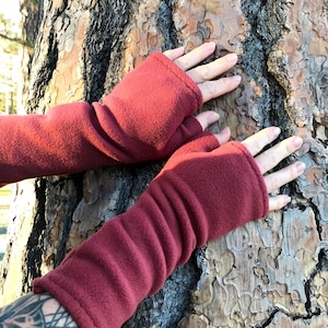 May include: Burgundy fleece fingerless gloves. The gloves are a deep red color and have a ribbed texture. The cuffs are finished with a simple hem. The gloves are shown against a tree trunk.