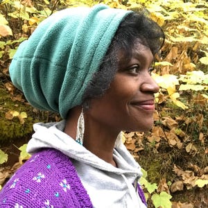 May include: A woman wearing a teal fleece beanie and a purple cardigan with white and teal accents. She is wearing long, beaded earrings.