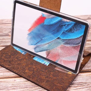 Könnte beinhalten: Eine braune Tablet-Hülle aus Leder mit floralem Prägemuster. Das Tablet wird mit einer transparenten Hülle angezeigt und zeigt ein farbenfrohes abstraktes Bild auf dem Bildschirm. Die Hülle hat Kartenfächer.