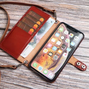 May include: A brown leather phone wallet case with a long shoulder strap, featuring multiple interior card slots, a cash compartment, and a secure snap button closure. The case holds a smartphone with a colorful digital display screen visible.