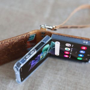 May include: A brown leather phone case with a floral embossed design, open to reveal a black smartphone with a clear protective case. The phone displays a colourful app screen. A matching leather strap is attached.