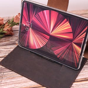 May include: A brown leather tablet case with a tablet inside, displaying a colourful abstract design. The tablet is in a clear case. The time on the screen reads 9:41. The case is open, resting on a wooden surface.