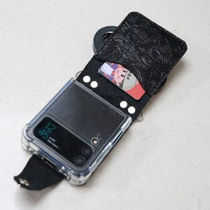 May include: Black leather phone case with a floral embossed design, featuring a clear phone case and a strap. The case holds a phone with a visible screen displaying the time. A card is visible inside the case.