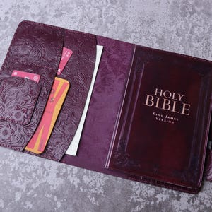 May include: Open burgundy leather Bible cover with embossed floral design. The cover holds a Bible with "Holy Bible" and "King James Version" text. Interior pockets hold cards and a white sheet of paper. A pen holder is visible on the right side.
