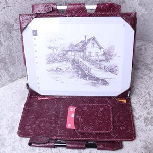 May include: A burgundy leather e-reader case with a floral embossed pattern. The case is open, displaying a digital screen with a black and white drawing of a house and bridge. The case has card slots and a pen holder.