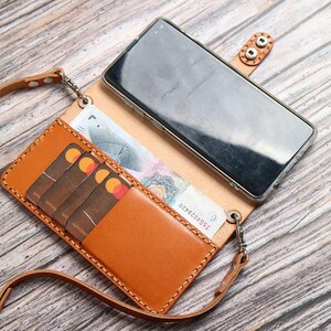 May include: A tan leather phone case with a strap, holding a smartphone, credit cards, and paper currency. The case has a snap closure and a stitched border. The strap is also made of tan leather.
