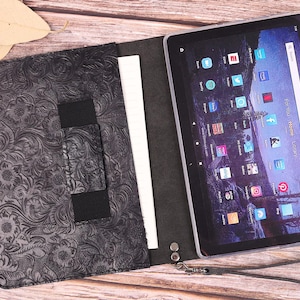 May include: Black tablet case with a floral embossed design, open to reveal a tablet with a colorful display. The case has a strap and a wrist loop. The tablet screen displays various app icons.