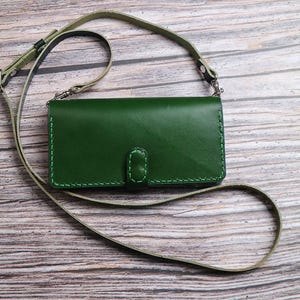 Pode incluir: Uma capa de telefone crossbody de couro verde com uma alça combinando. A capa retangular tem uma borda costurada e um fecho de aba. A alça é de cor verde oliva clara. A capa está sobre uma superfície de madeira.