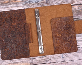 May include: A brown leather binder with a floral embossed design. The binder has a metal ring mechanism and a flap closure.