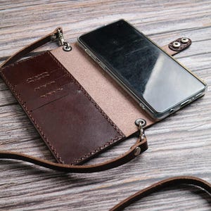 May include: A brown leather phone case with a strap, open to reveal a phone and card slots. The case is made of dark brown leather with a lighter brown interior and a snap closure. The phone is in a clear case.