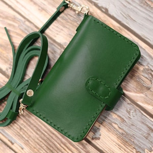 May include: A forest green leather phone case with a matching strap. The case has a secure flap closure and is stitched with a contrasting thread. The strap is attached with silver-toned hardware. A stylish and functional accessory.