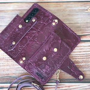 May include: A deep purple leather phone case with a floral embossed design. The case has a wallet-style design with multiple compartments and a strap. Gold-colored hardware accents the case, including snap closures and a strap attachment. The case is on a wooden surface.