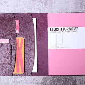 Könnte beinhalten: Geöffnetes lila Lederjournal mit geprägtem floralem Design. Im Inneren befindet sich ein rosa Notizbuch mit dem Text "LEUCHTTURM1917". Es gibt Kartenfächer und eine Stifthalterung.