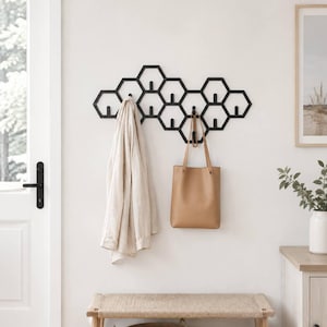 May include: A matte black wall-mounted coat rack featuring a geometric honeycomb pattern made of interconnected hexagonal frames. The rack includes multiple small vertical hooks positioned within the centre of several hexagons for hanging items.