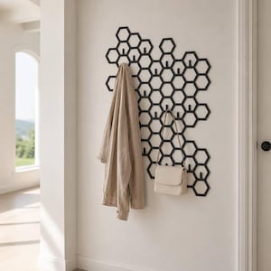 May include: A black metal wall-mounted coat rack featuring a geometric honeycomb pattern with integrated hooks. A beige button-down shirt and a small cream-coloured shoulder bag are hanging from the hooks of this modular hexagonal storage unit.