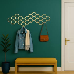 Op de afbeelding: Een goudkleurige, honingraatvormige wandkapstok met meerdere haken. Een spijkerjasje en sjaal hangen aan de kapstok, met een bruine handtas. Een gele bank en een patroonkleed maken de entree compleet.