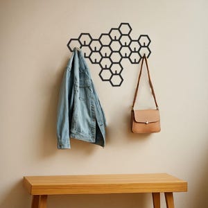 May include: A black metal honeycomb wall-mounted coat rack with a light blue denim jacket and a tan purse hanging from it. A wooden bench sits below the rack. The wall is a light beige color.