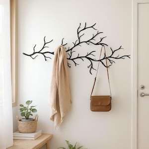 May include: A black metal wall-mounted coat rack in the shape of tree branches. A tan scarf and a brown purse hang from the branches. A small bench with a plant in a basket and a potted fern are in the foreground.