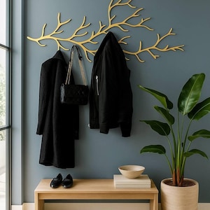 Puede incluir: Un perchero de pared de color dorado con forma de rama de árbol. El perchero cuenta con múltiples ganchos para colgar objetos y está montado horizontalmente en la pared. Está fabricado en metal con un acabado liso y mate.